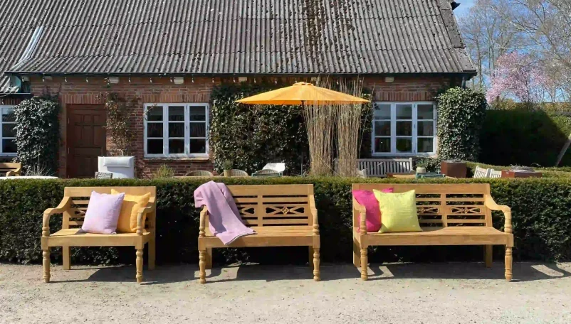 Holzbänke mit Kissen und einem Sonnenschirm vor einem Haus und Hecken.