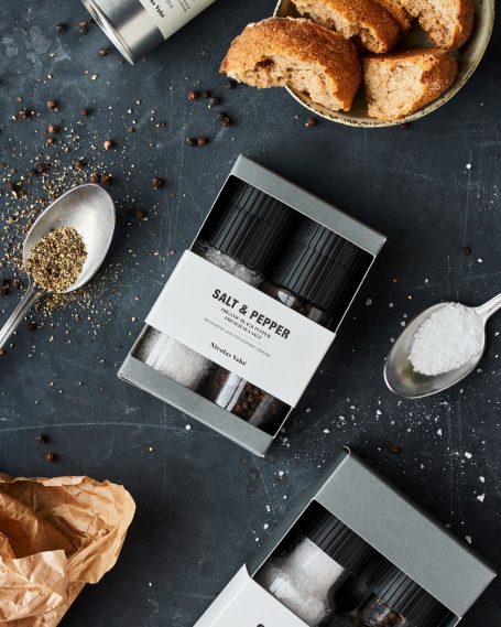 Geschenkset mit Gewürzen, Salz und Pfeffer in eleganter Verpackung, umgeben von Brotkrümeln.