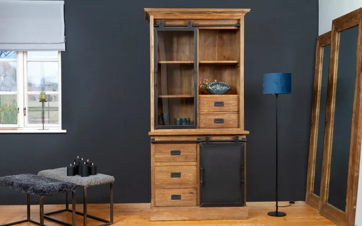 Holzregal mit Glasfront, Schubladen und moderner Stehlampe neben einem Sofa.