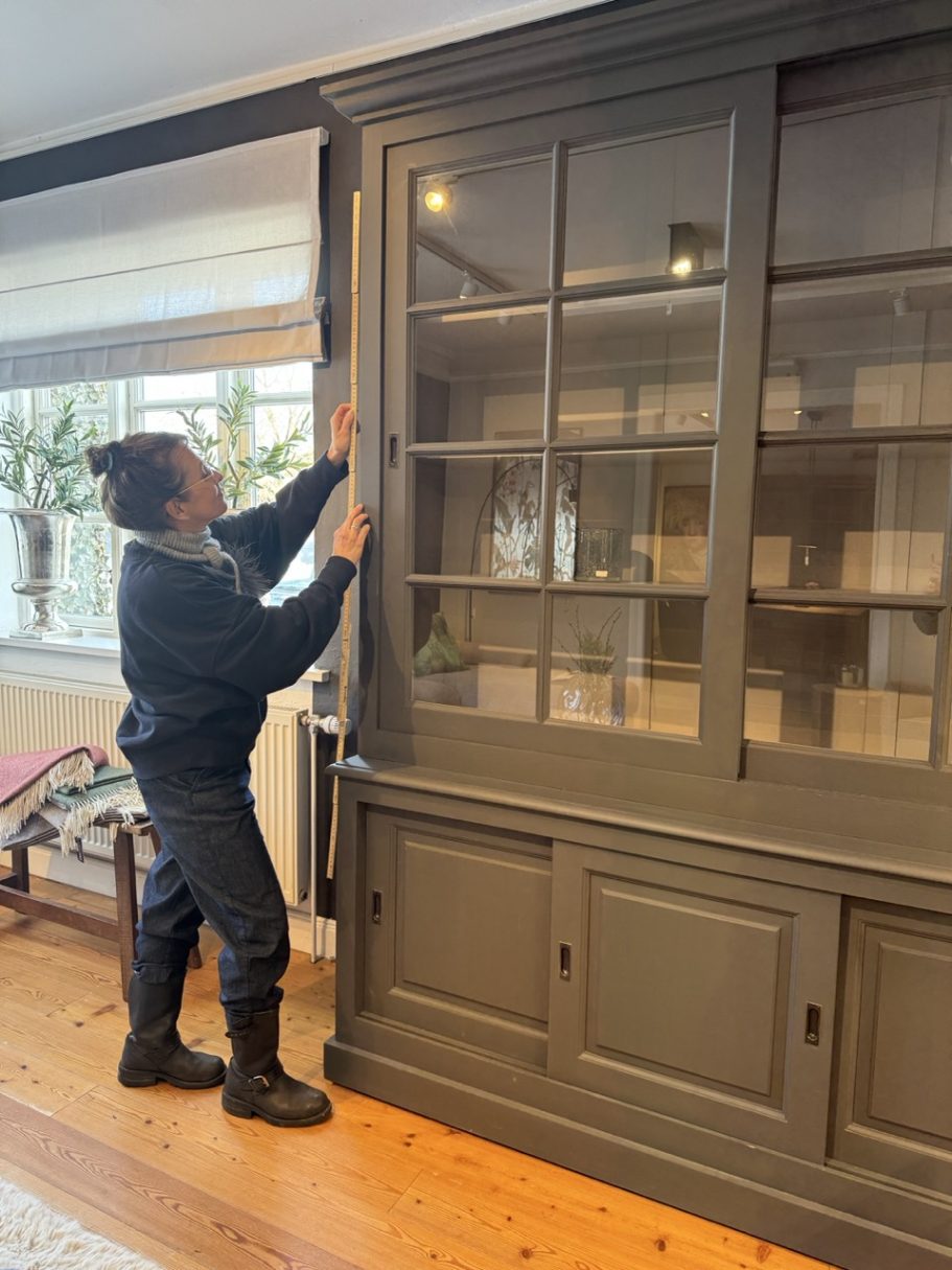 Eine Person in schwarzer Kleidung steht vor einem grauen Schrank und misst ihn aus.