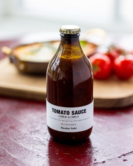 Glasflasche mit Tomatensoße, umgeben von frischen Tomaten und einem Gericht im Hintergrund.